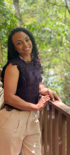 Ana Nascimento em pé, sorrindo, apoiada em um guarda-corpo de madeira, com vegetação ao fundo.